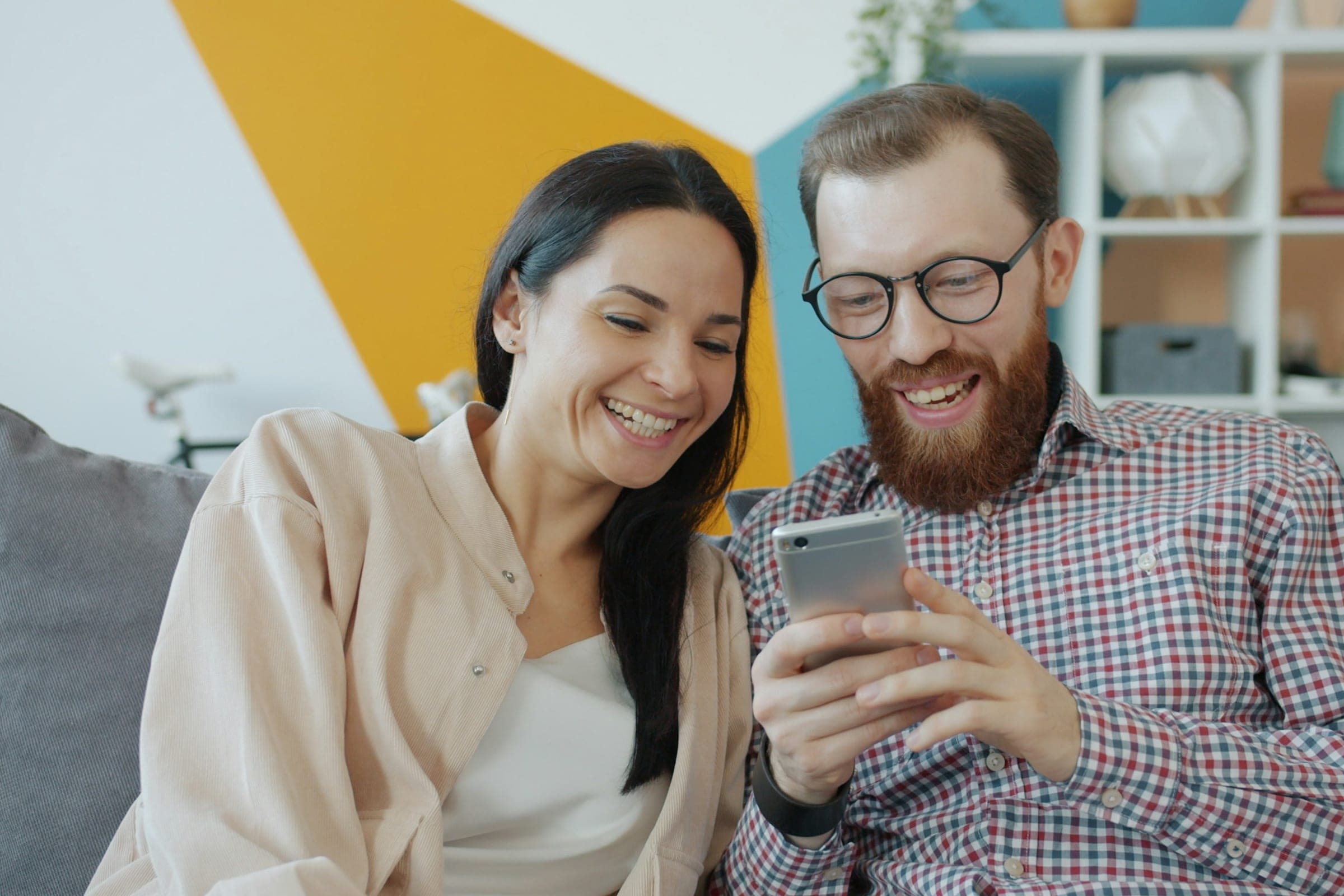 Two friends laughing together on a sofa as they look at offers on a phone