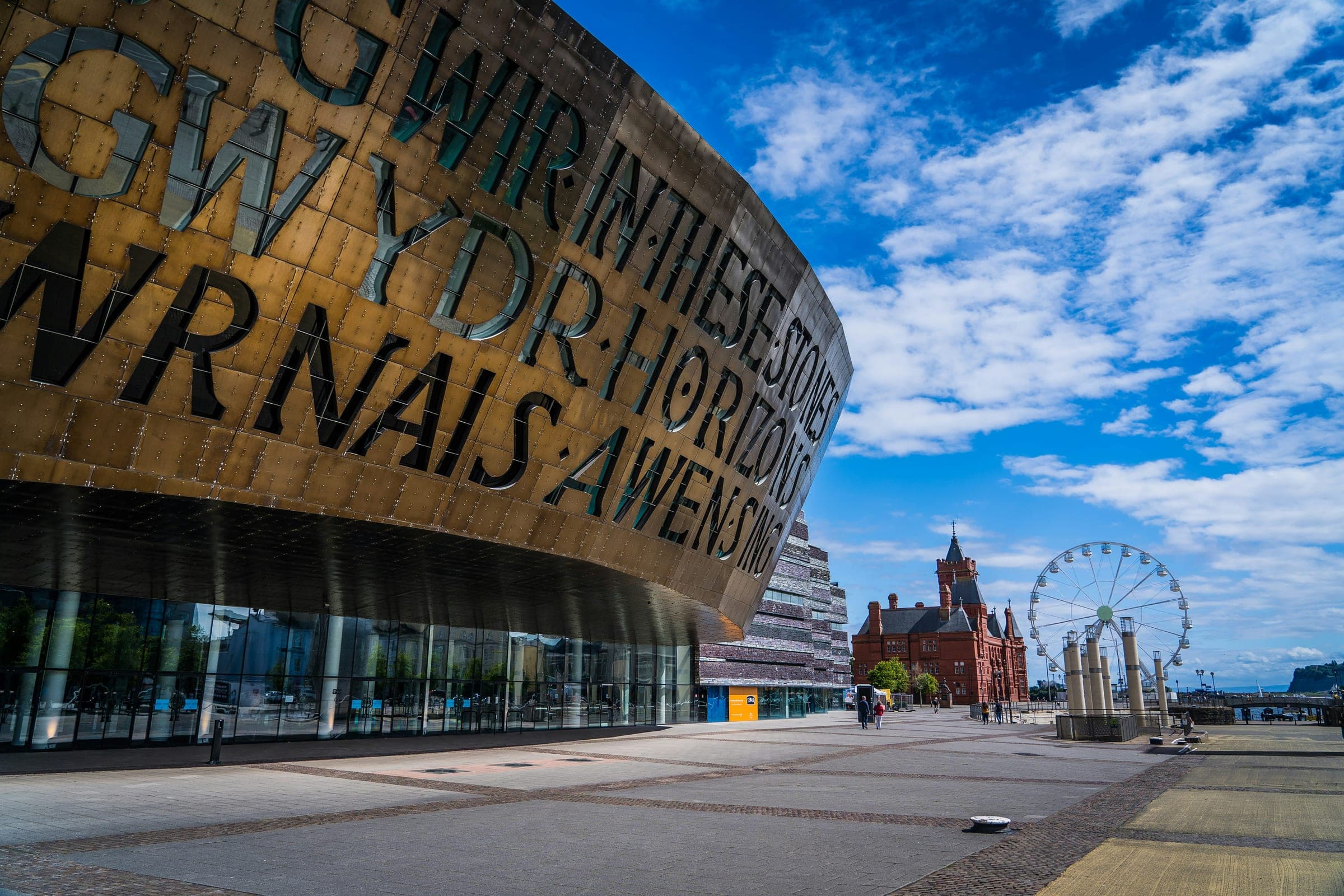 The Wales Millennium Centre in Cardiff Bay under a blue sky, with the Pierhead Building and the big wheel beyond