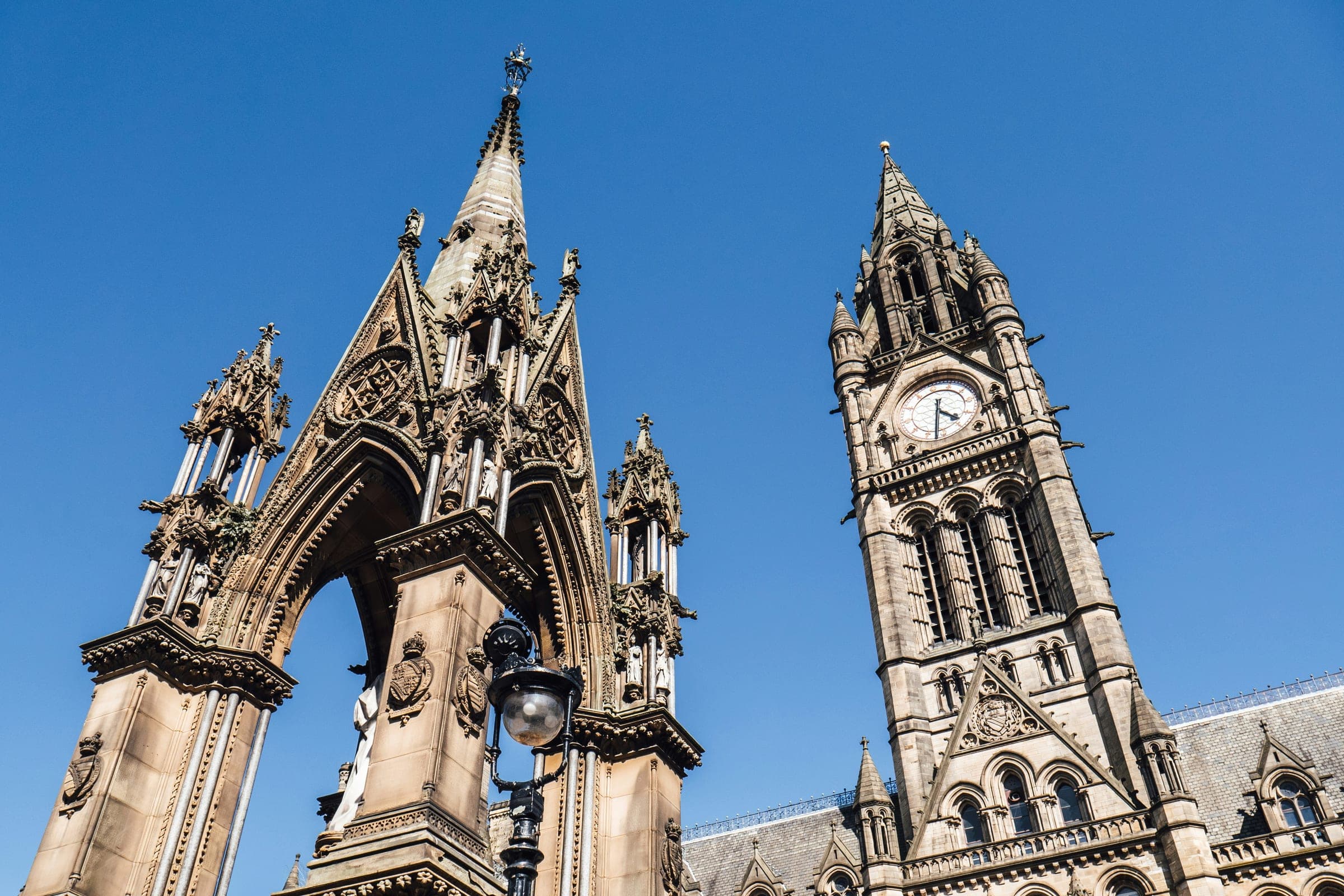 The Albert Memorial and Manchester Town Hall’s clock tower in Albert Square under a blue sky