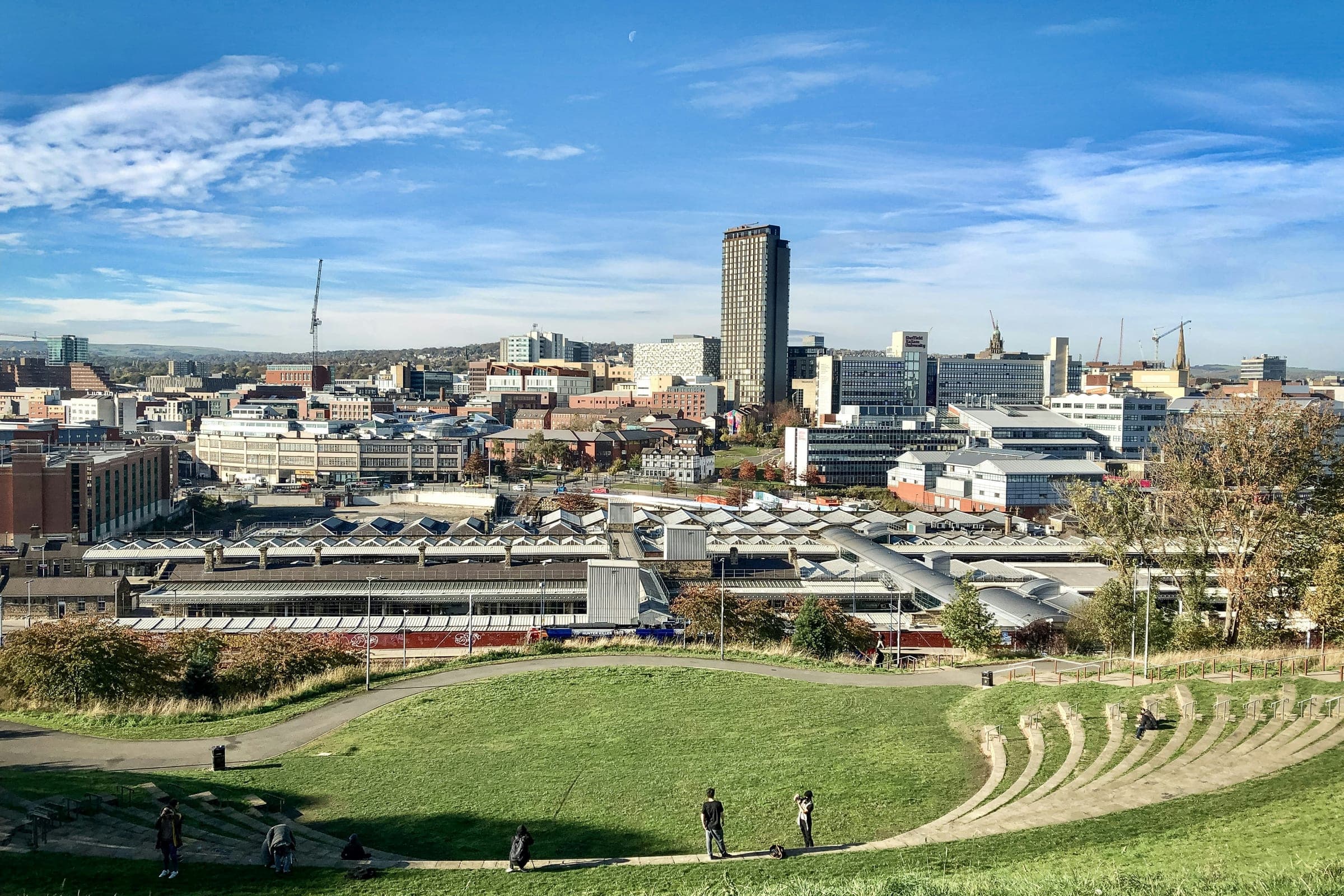 People walking in the sunshine on the hillside above Sheffield, with the city centre and the station below