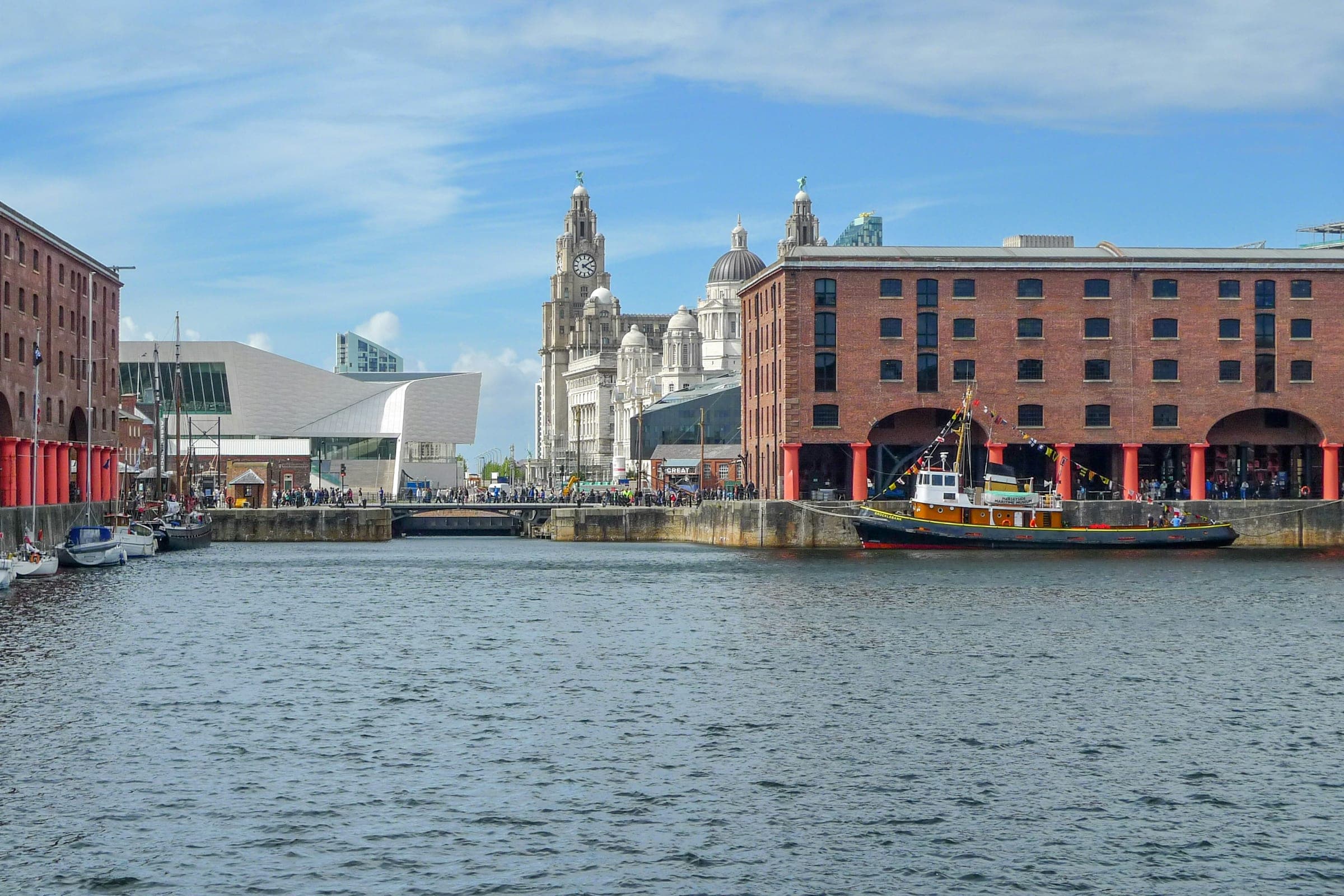 Crowds on the quayside at the Royal Albert Dock on a sunny day, with the Three Graces behind